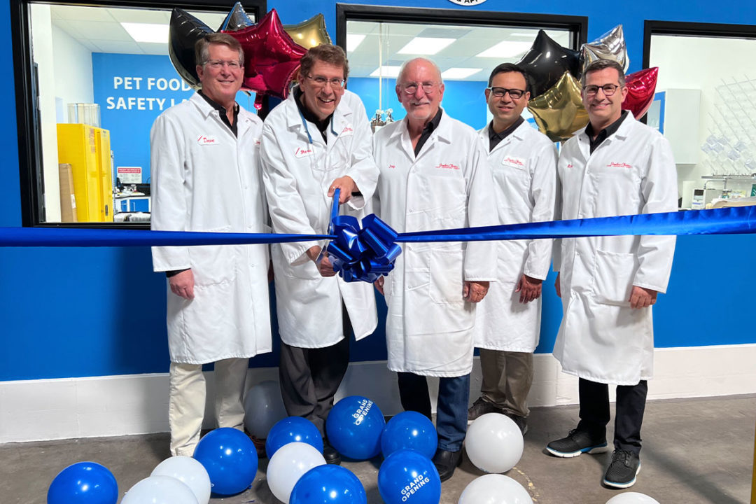 Five men in white lab coats surrounded by blue and white balloons while one man cuts a large blue ribbon.