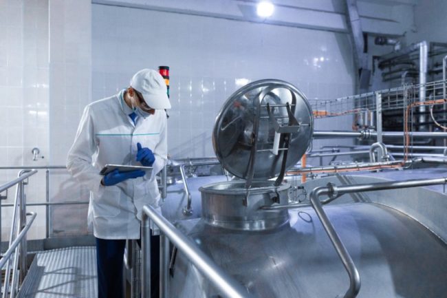 Man in white lab coat and hard hat holding clip board examines a dairy milk tank.