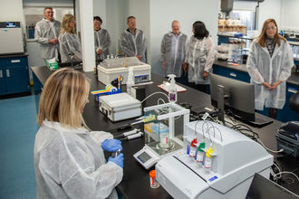 Men and women wearing disposable lab frocks and safety glasses stand around a table filled with laboratory equipment. A woman wearing blue safety gloves is opening a bottle.